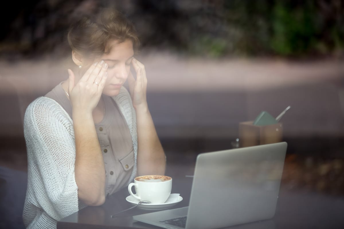 Stressed woman sitting at a café table with a laptop, holding her head from emotional exhaustion.